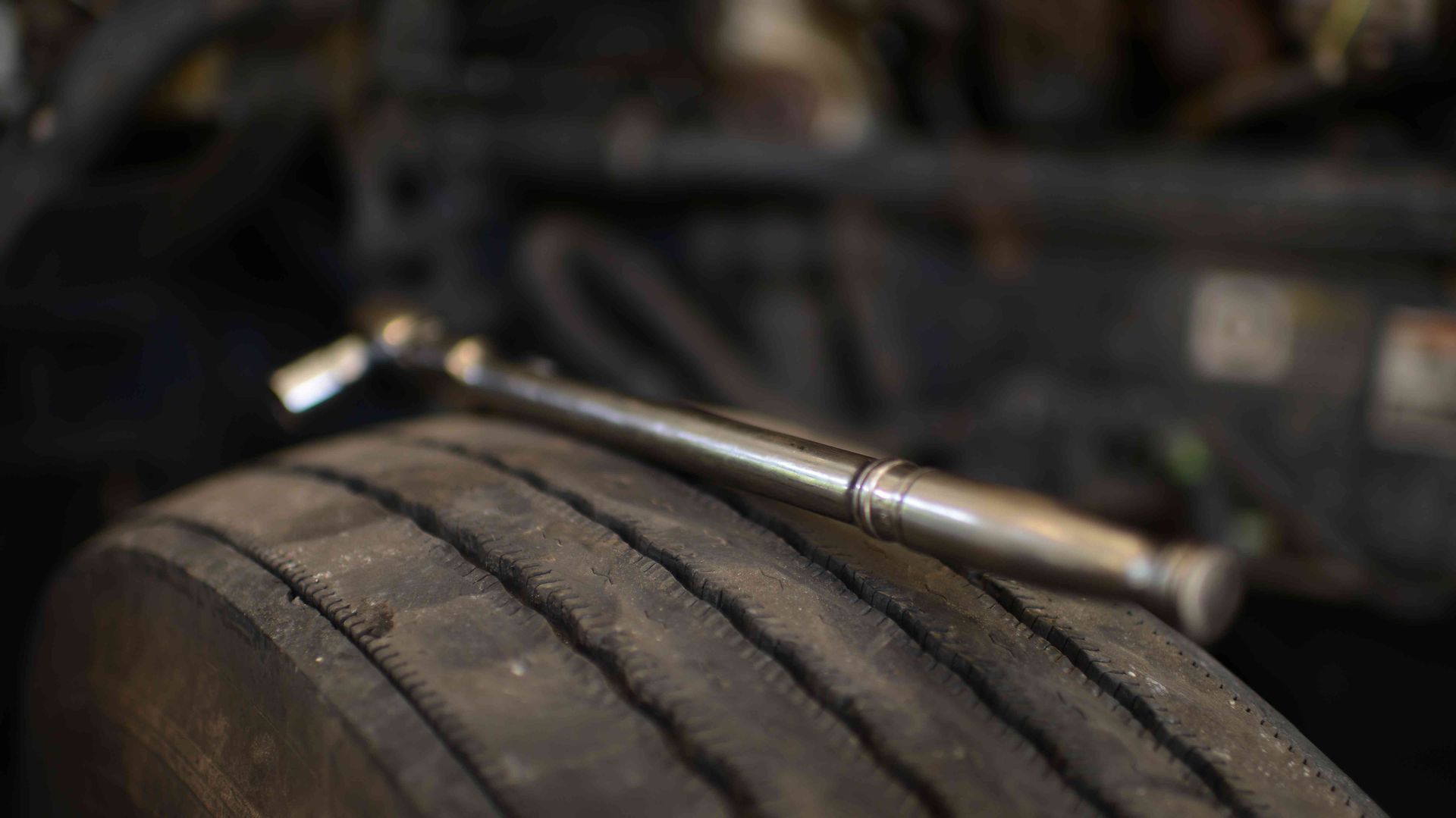 Silver tire tread depth gauge resting on a worn black tire in a dim garage.