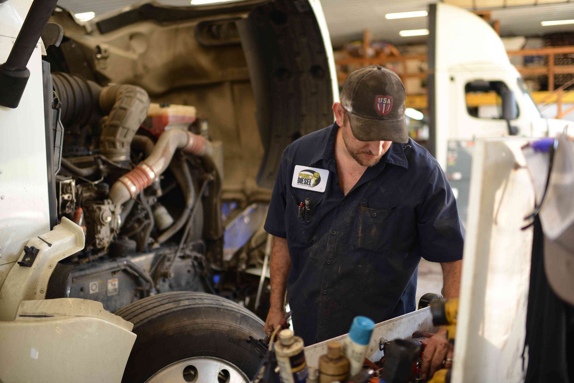 Mechanic in navy uniform inspecting a truck engine in a garage, with tools nearby.