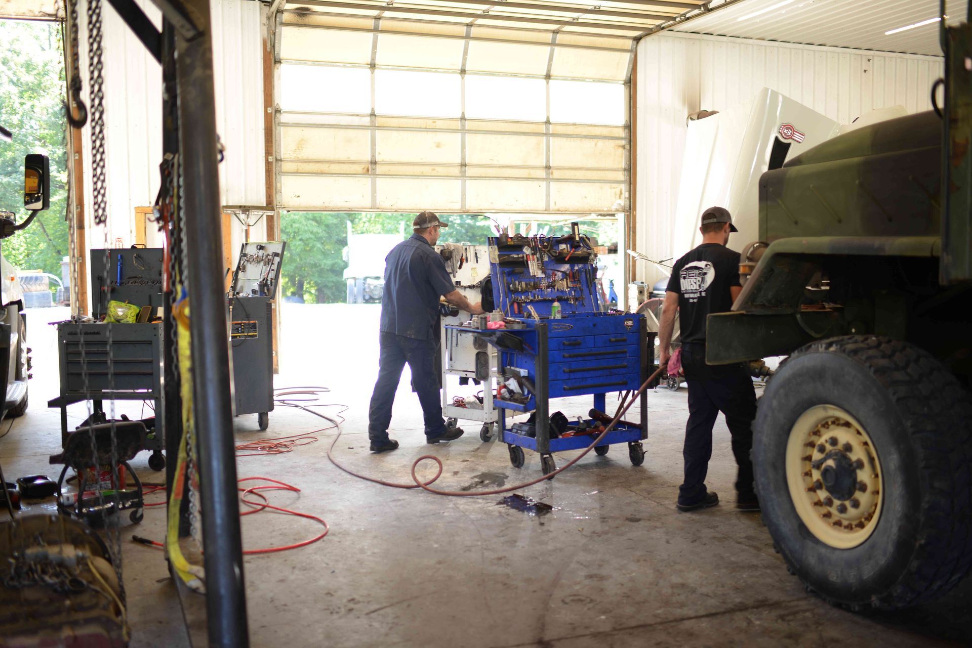 Two mechanics work in a garage beside a large truck and blue engine equipment.