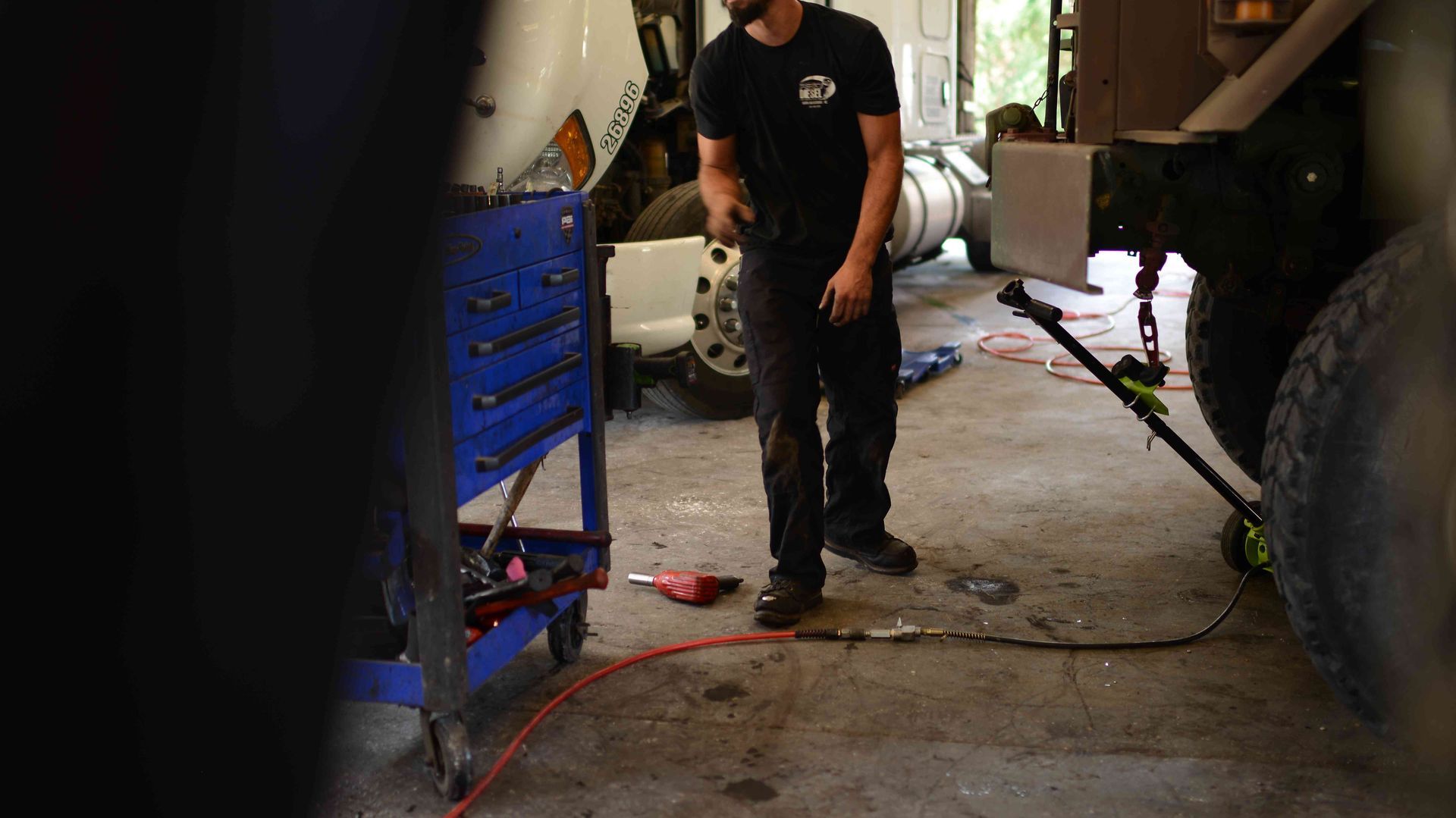 Mechanic standing in a garage beside a blue tool cart and large vehicle tire, with a red air hose on the floor