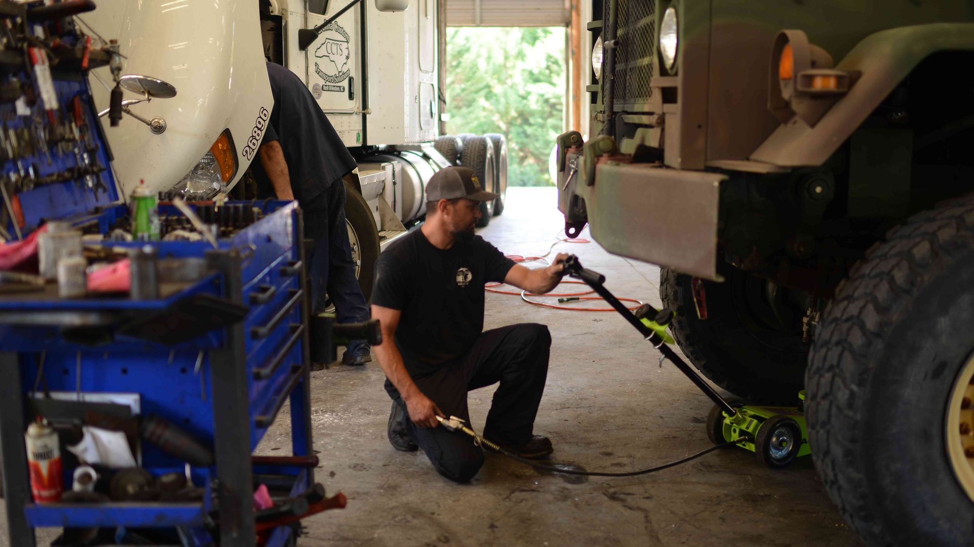 Mechanic kneeling beside a large truck in a garage, inspecting undercarriage equipment.