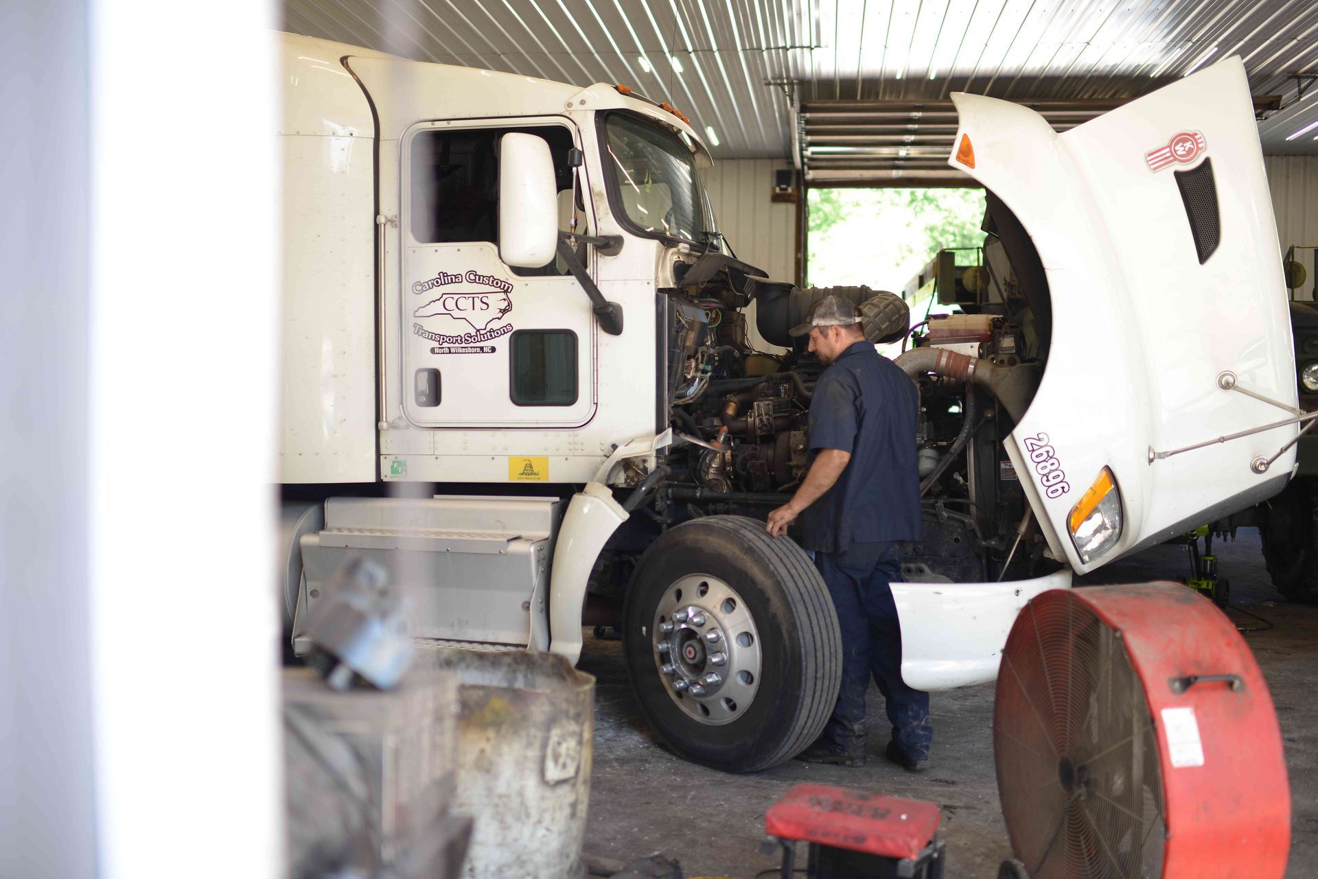 White semi-truck being repaired inside a garage, with a mechanic working on the open engine.