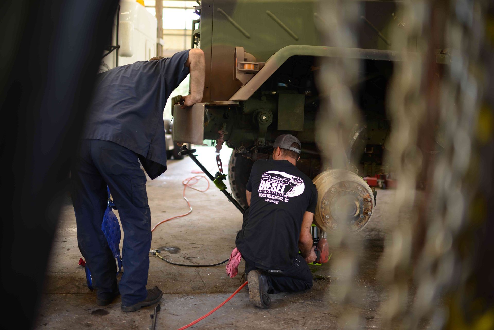 Mechanics working under a large vehicle, with one kneeling and another standing nearby