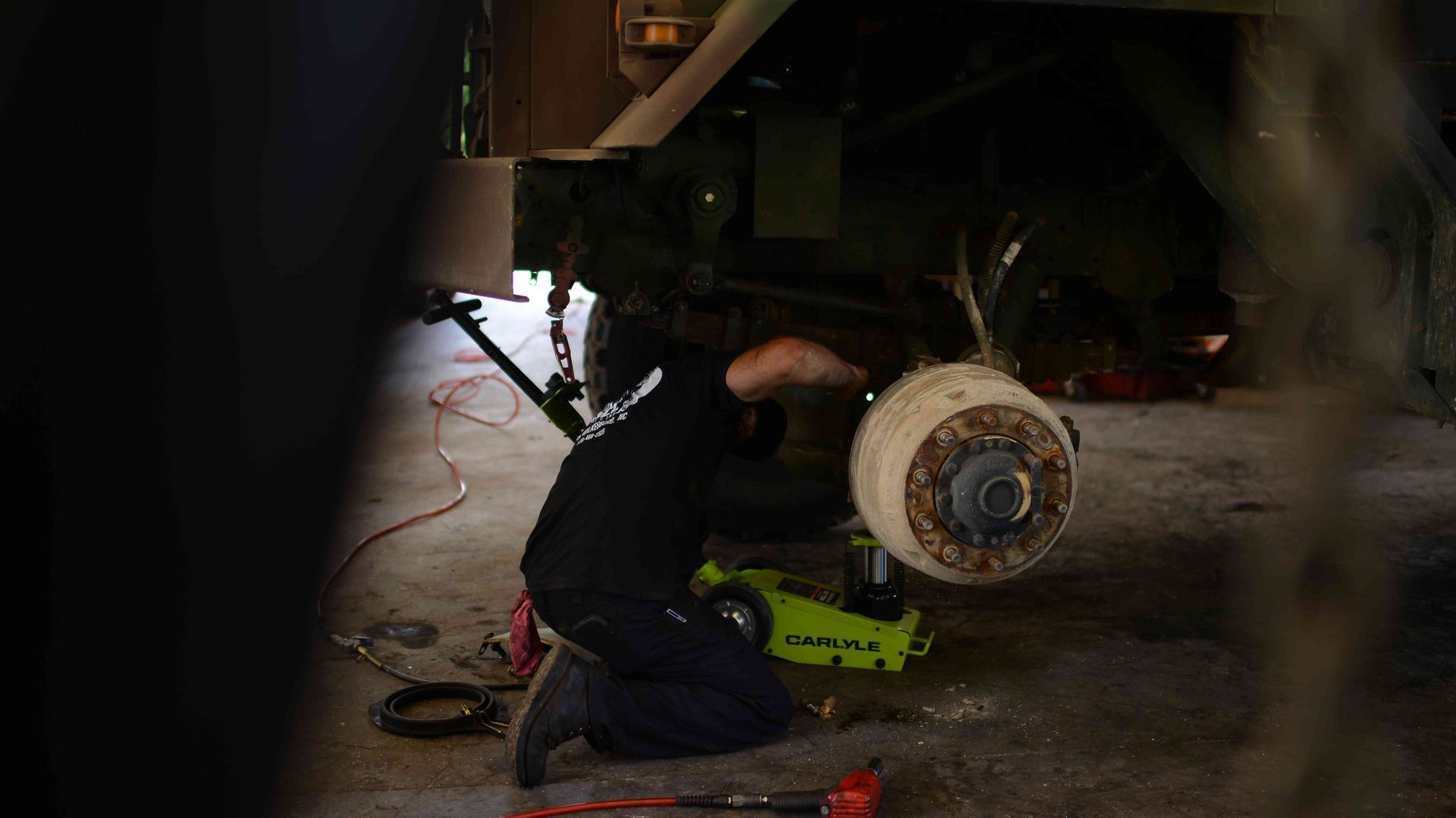 Mechanic kneeling under a vehicle, using a green power tool near a brake assembly in a dim garage.