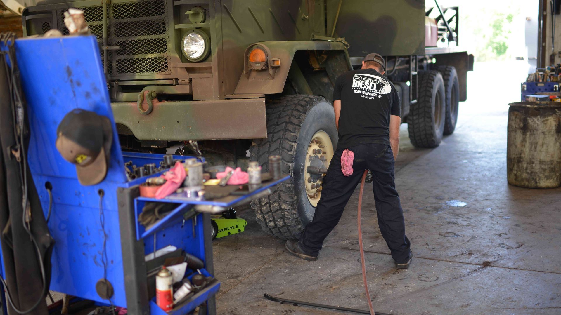 Mechanic working beside a large military truck in a garage, with tools and equipment nearby
