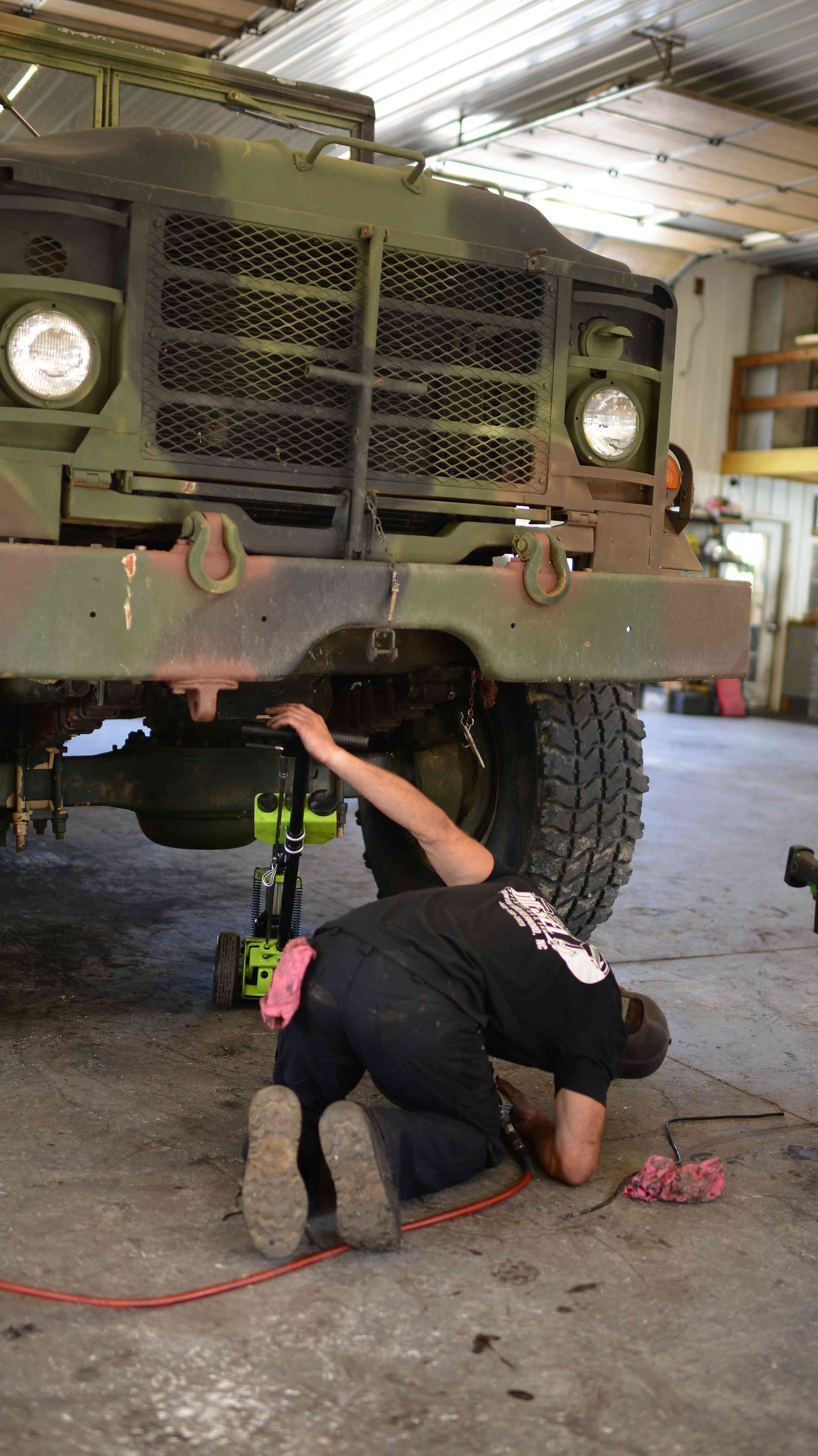 Person works under a lifted military truck in a garage, using a tool on the undercarriage.