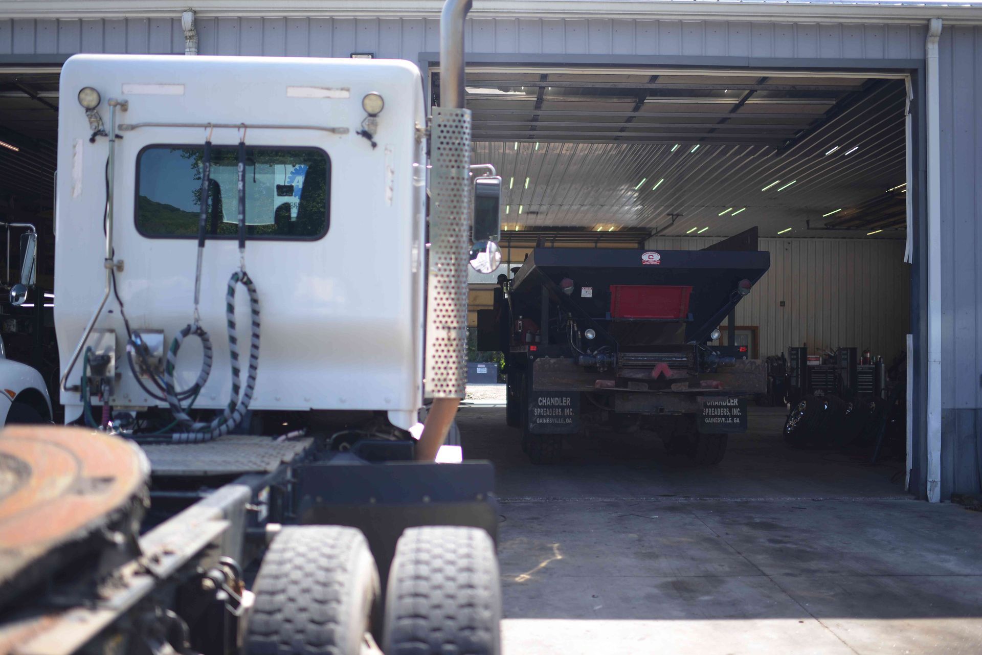 White truck and dump trailer in a garage bay, with a large off-road tire in the foreground.