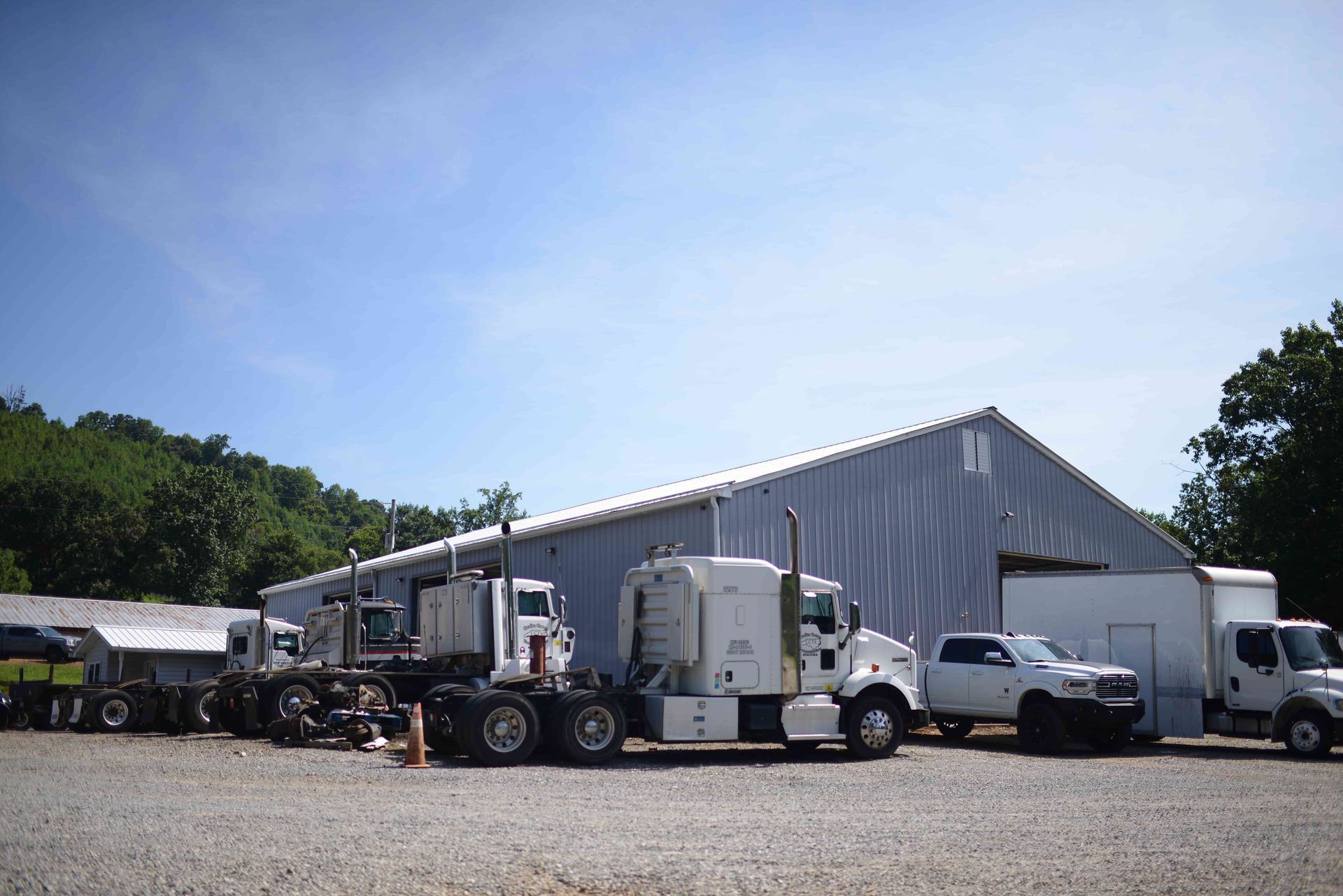 Parked semi-trucks and trailers beside a large gray warehouse on a sunny gravel lot.
