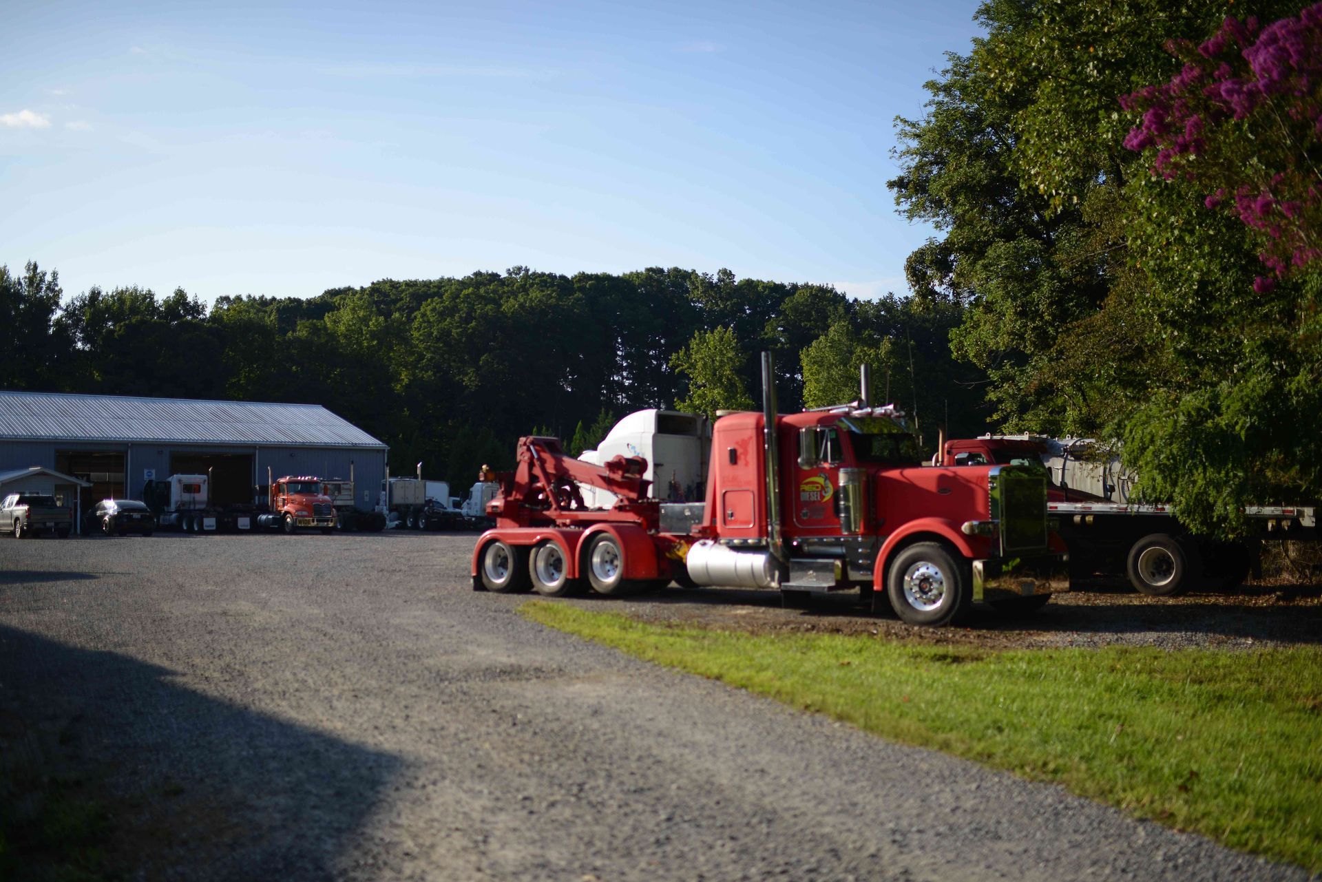 Red tow truck parked on a gravel lot beside a garage, with a trailer and trees in the background.