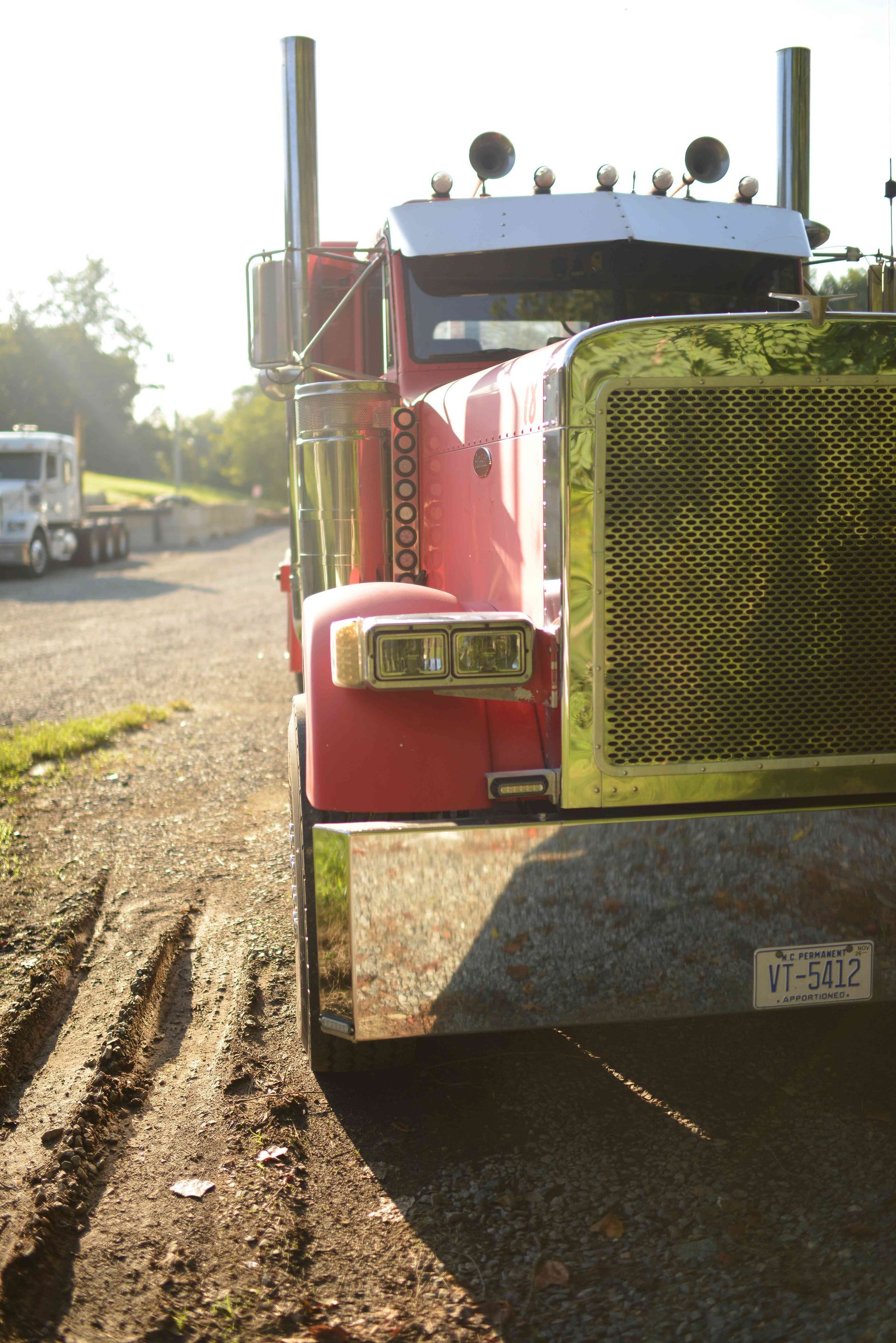 Red semi-truck parked on a dirt road in bright daylight, front grille facing the camera.