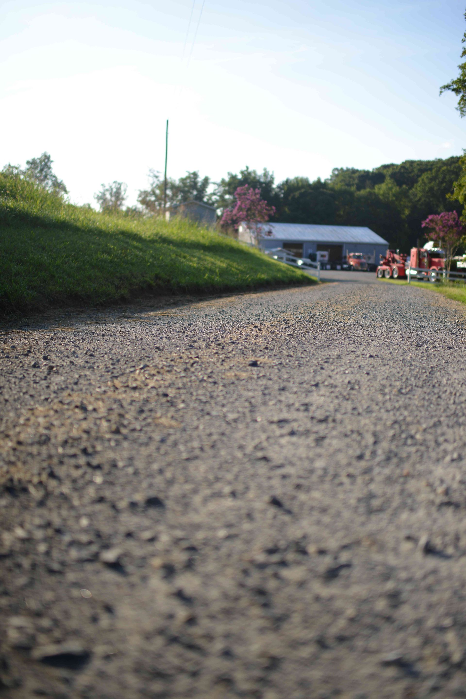 Low-angle view of a gravel path beside a green embankment, leading to houses and trees in the distance.