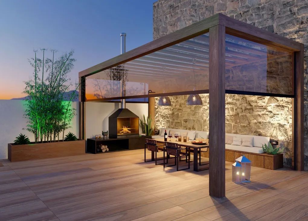 Outdoor dining area with a wooden pergola, fireplace, and stone wall, at dusk.