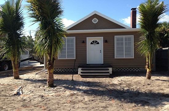 Small brown house with white trim and palm trees in a sandy yard