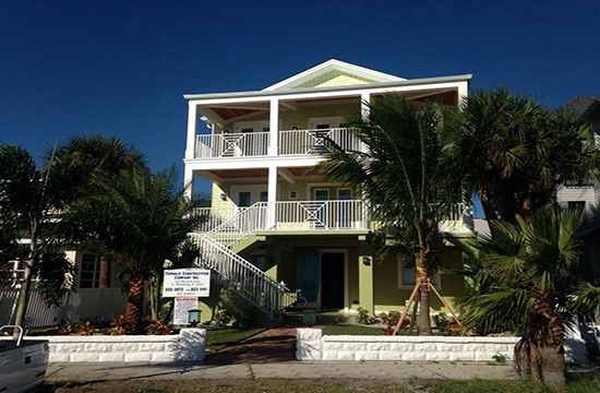 Two-story white beach house with palm trees and a front staircase under a blue sky