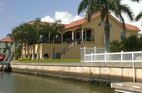 Canal-side house with palm trees, red roof, and white fence along the water