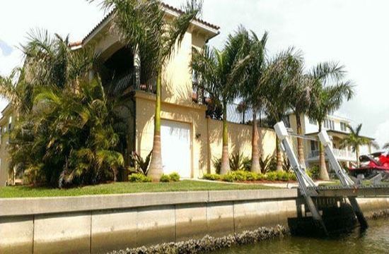 Waterfront home with palm trees and a dock beside a canal under a bright sky