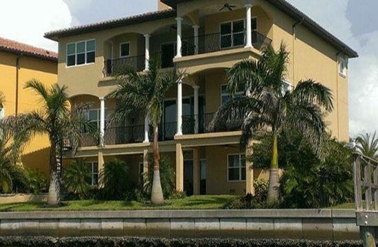 Three-story yellow apartment building with balconies, palm trees, and a canal in front