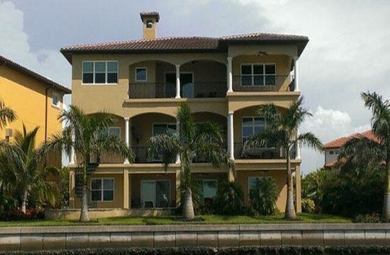 Two-story cream house with arched balconies, palm trees, and a driveway in front