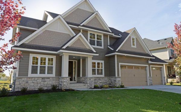 Two-story beige house with stone accents, beige garage doors, and a green lawn under a blue sky.