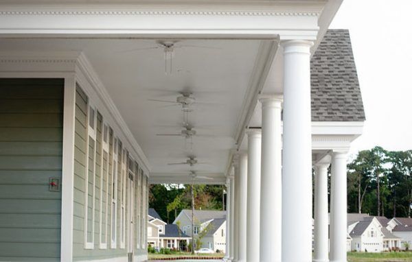 A long porch with white columns and ceiling fans; green siding; houses in the background.