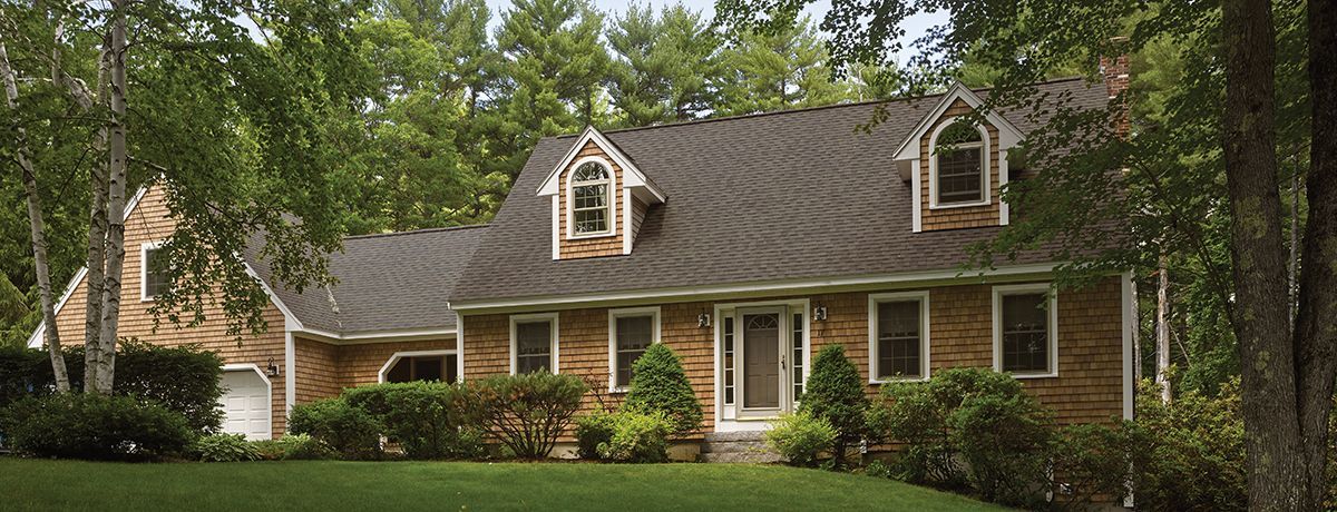 House with brown shingles, brick walls, and arched windows in a tree-lined yard.