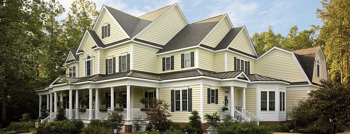 Large yellow house with multiple gables and a wrap-around porch, set among trees on a sunny day.