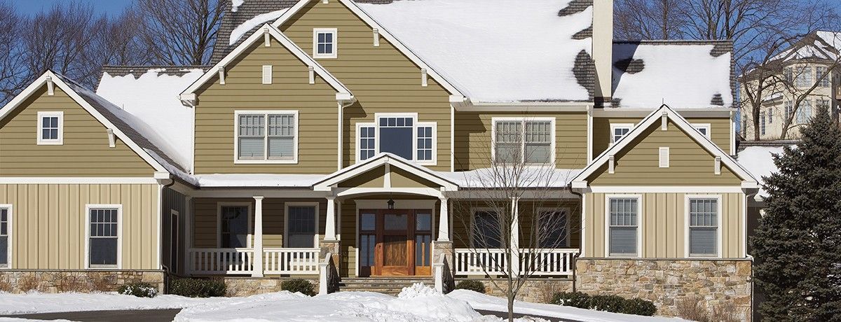 A two-story house covered in snow in a winter setting, with a brown wooden door and light-colored siding.