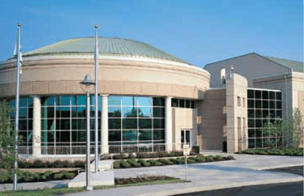 Large beige building with a round section, many windows, and a green dome on a sunny day.