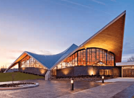 Modern building with soaring, angular roof; large glass windows; evening setting.
