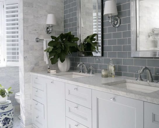 White bathroom with dual sinks, gray tiled wall, mirror, and large plant.