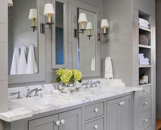 Gray bathroom with marble countertop, double sinks, mirrors, sconces, and built-in shelving with towels.