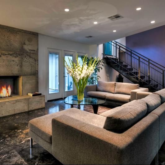 Modern living room with fireplace, couches, glass table, stairs, and large floral arrangement.