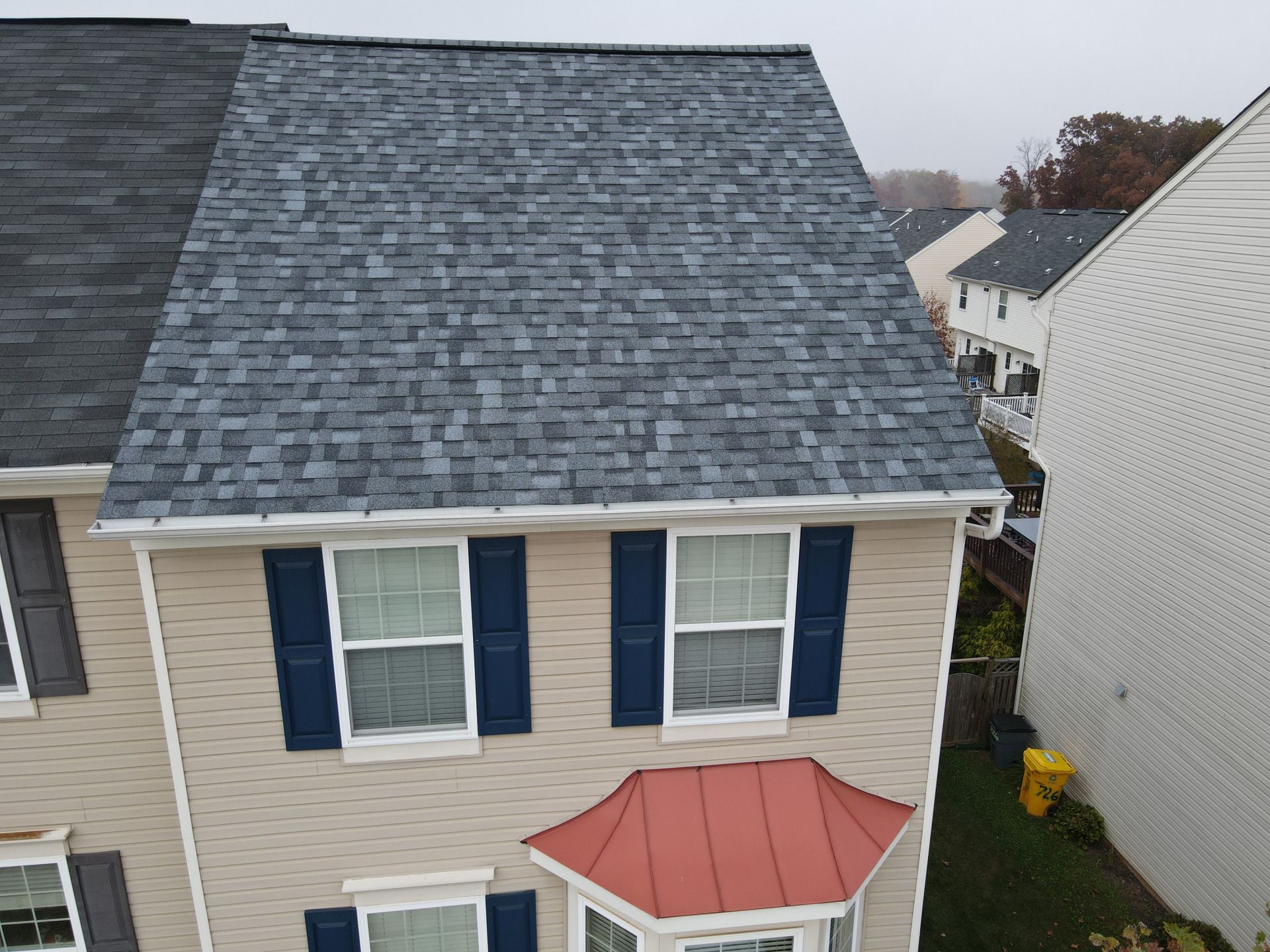 Beige house with blue shutters and a gray shingled roof. A red metal roof accents a bay window.