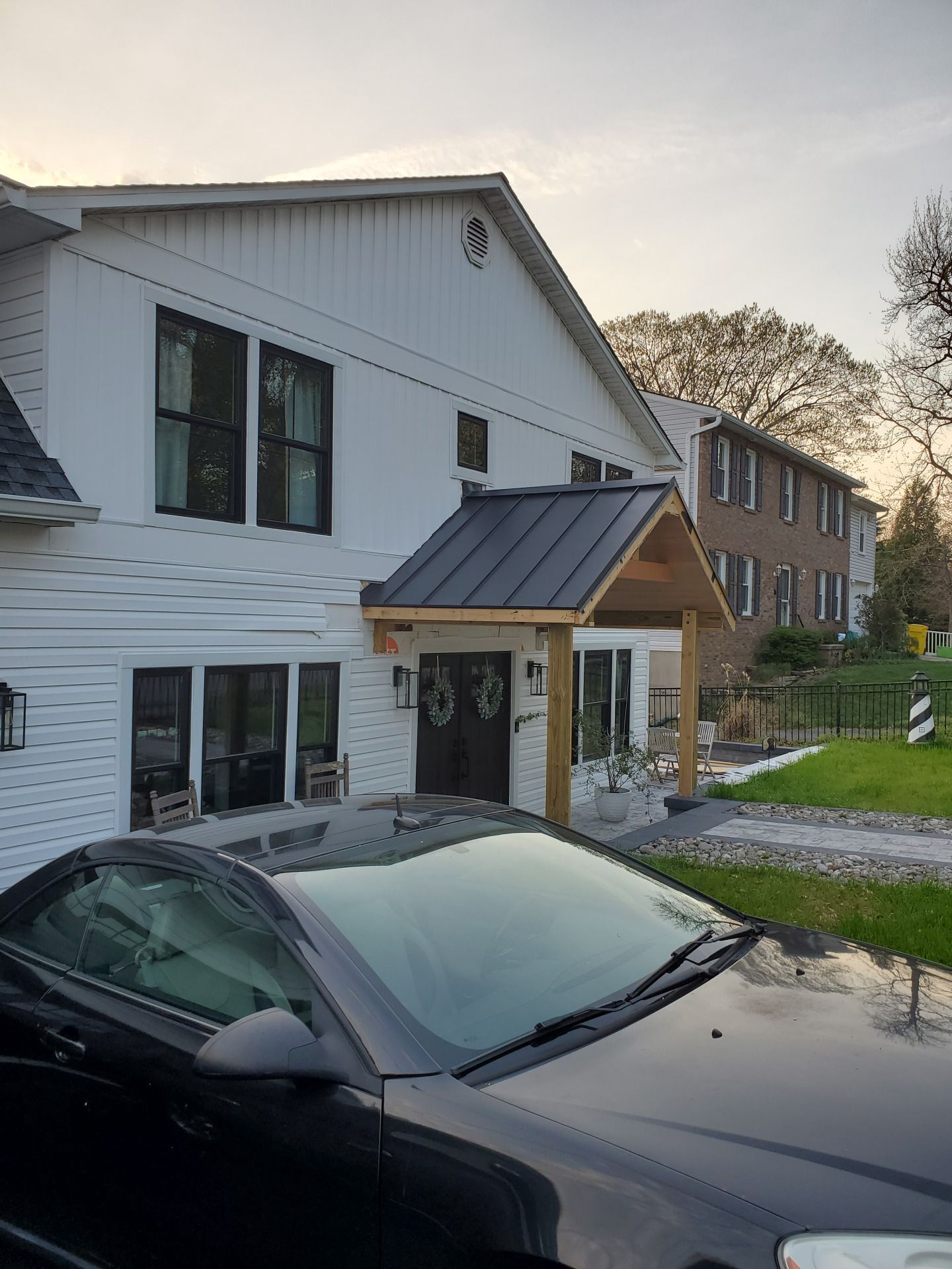 White house with black trim, a black car parked in front, and a brick building in the background.