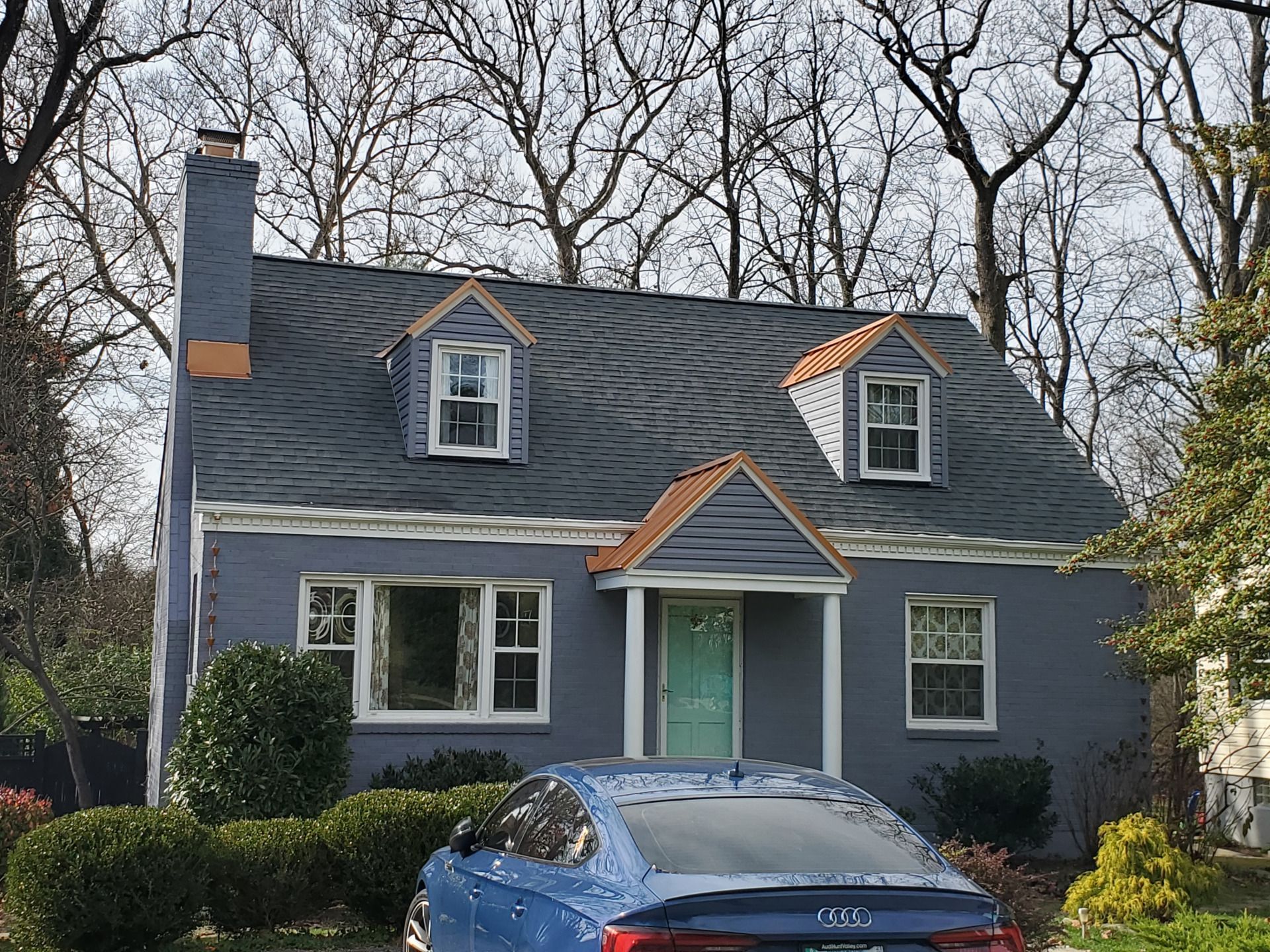 Blue house with dormers, teal door, car parked out front. Trees in the background.