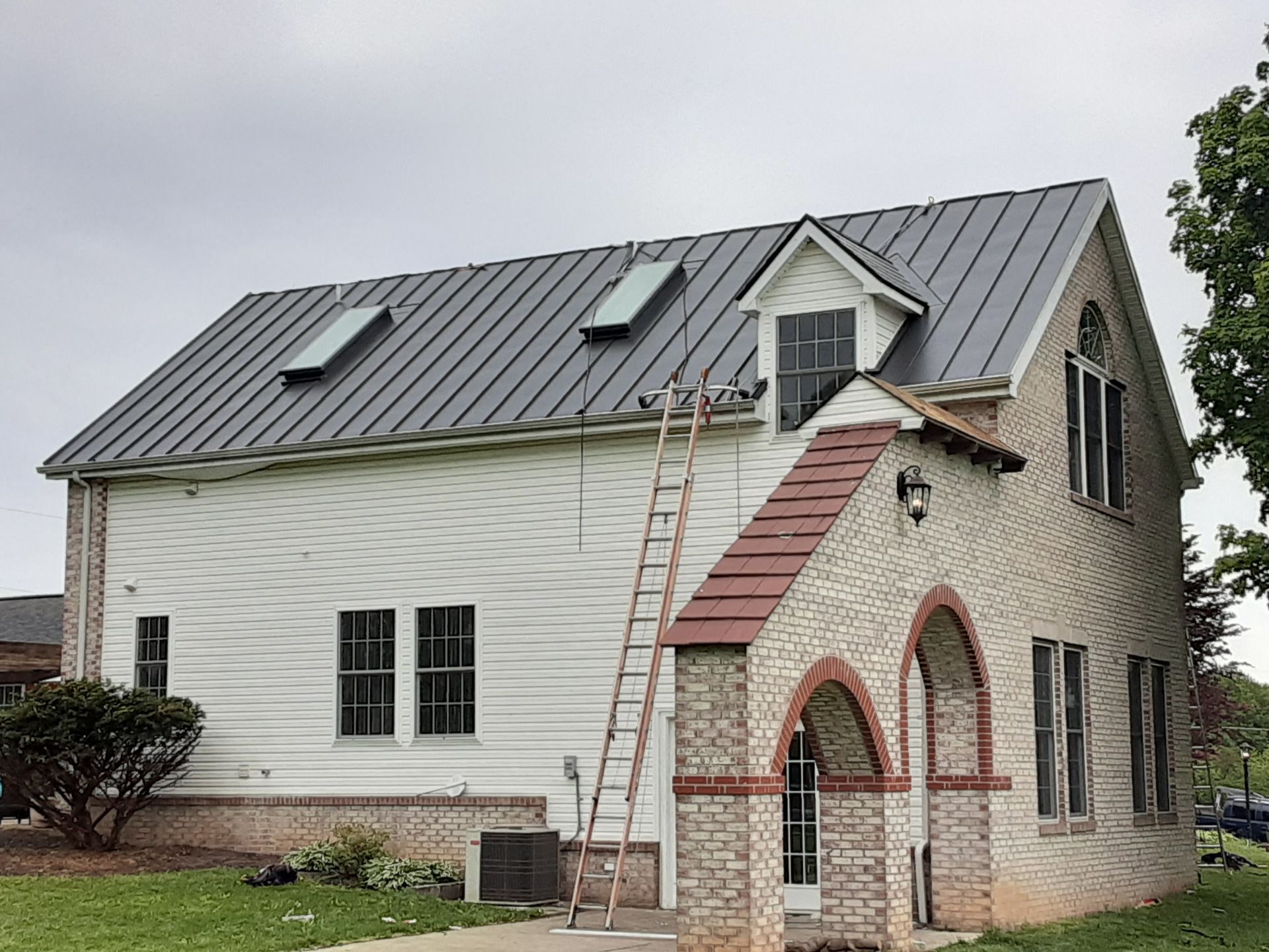 House with gray metal roof, white siding, brick arches, and a ladder.