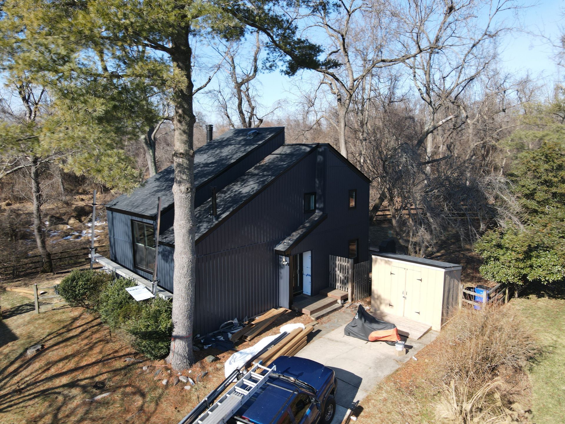 Black house with dark roof nestled in wooded area; driveway, trees, and blue truck.
