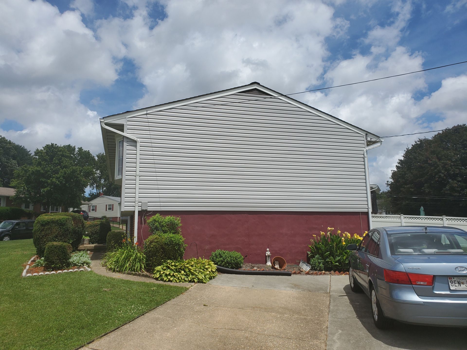 Side of a house with white siding, red brick base, and blue car in driveway, under a cloudy sky.