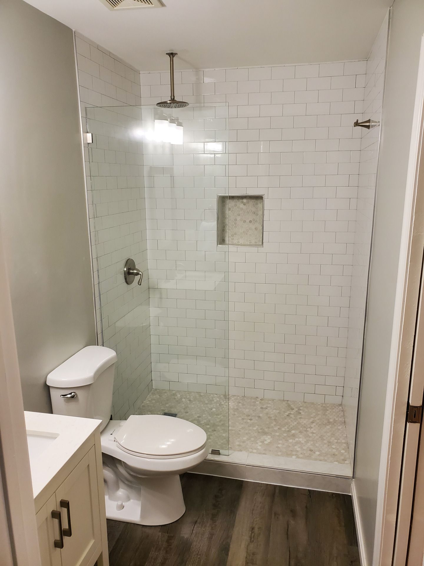 Bathroom with glass shower, white subway tile, toilet, vanity, and wood-look floor.