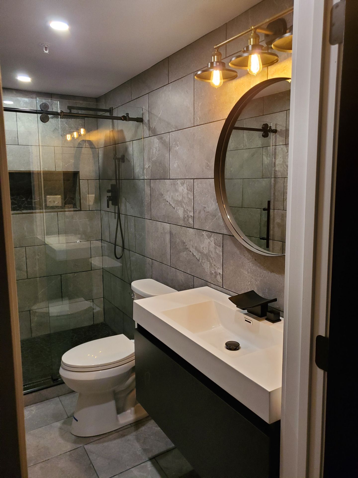 Modern bathroom with gray tile walls, black fixtures, and a round mirror.