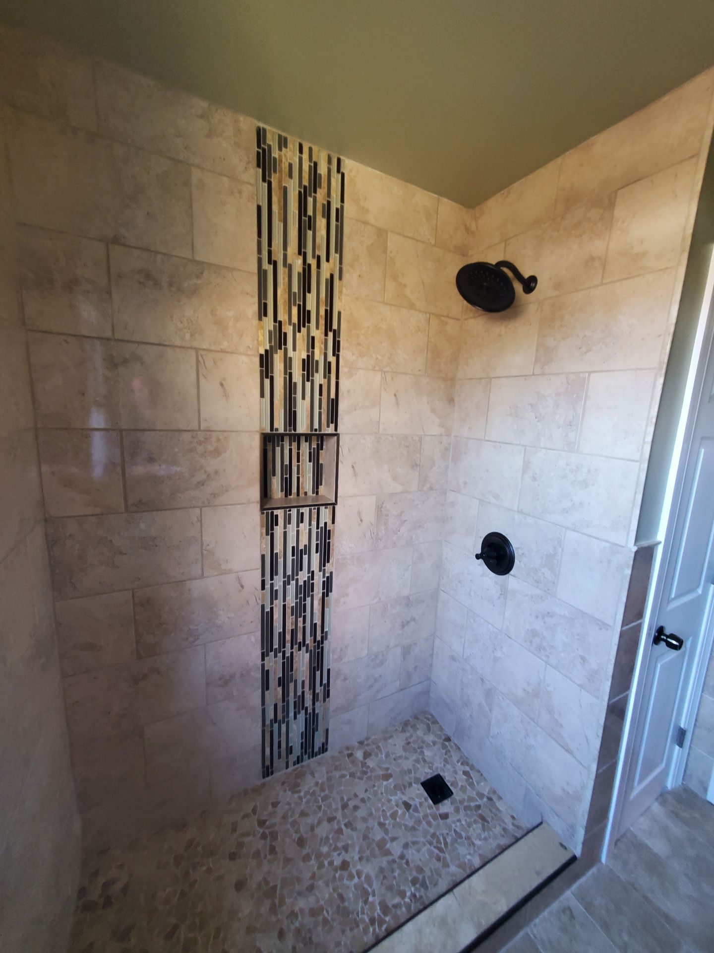 Shower with light beige tile, black fixtures, pebble floor, and a decorative mosaic.