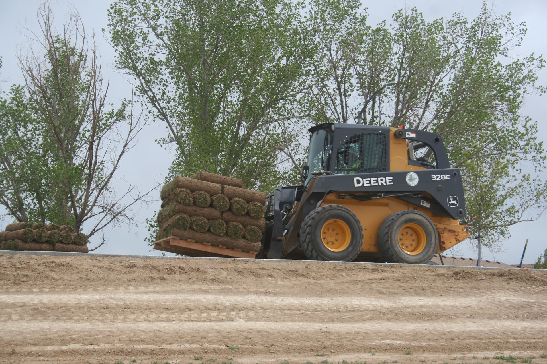 A John Deere skid steer carrying a pallet of rolled sod, in a dirt field with trees.