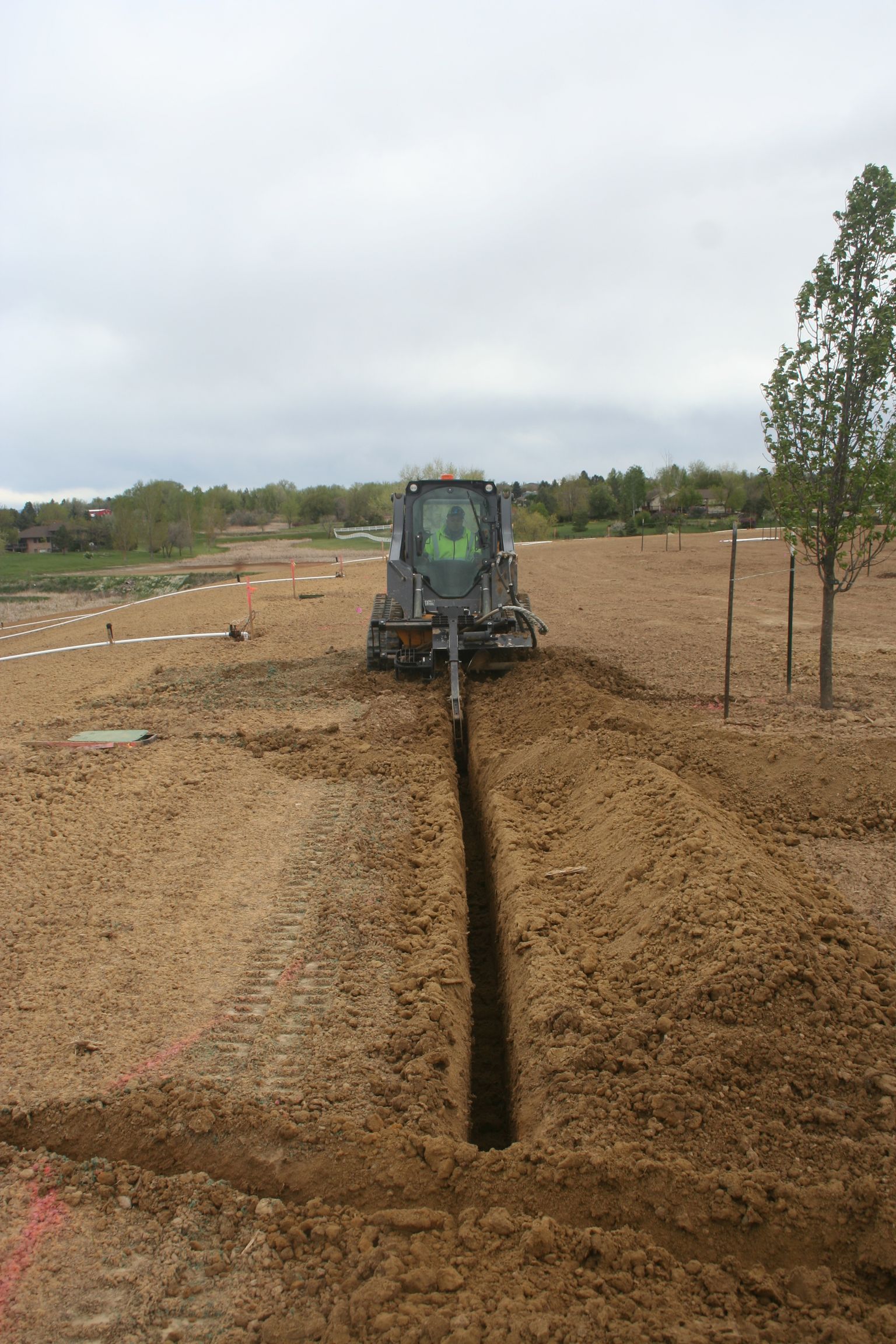 Tractor digging a trench in a dirt field under an overcast sky.