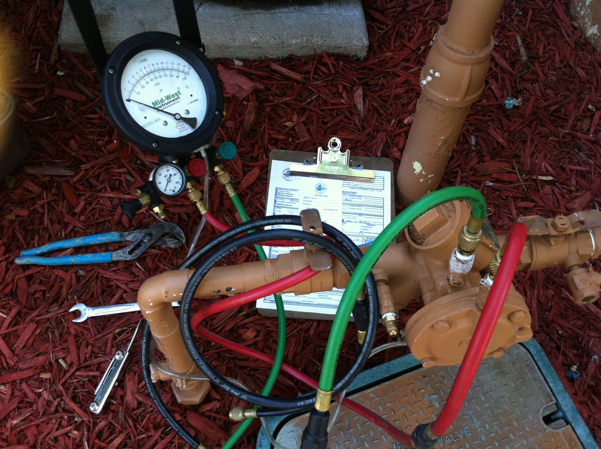 Pressure gauge and tools on top of a sprinkler system valve in a yard with red mulch.