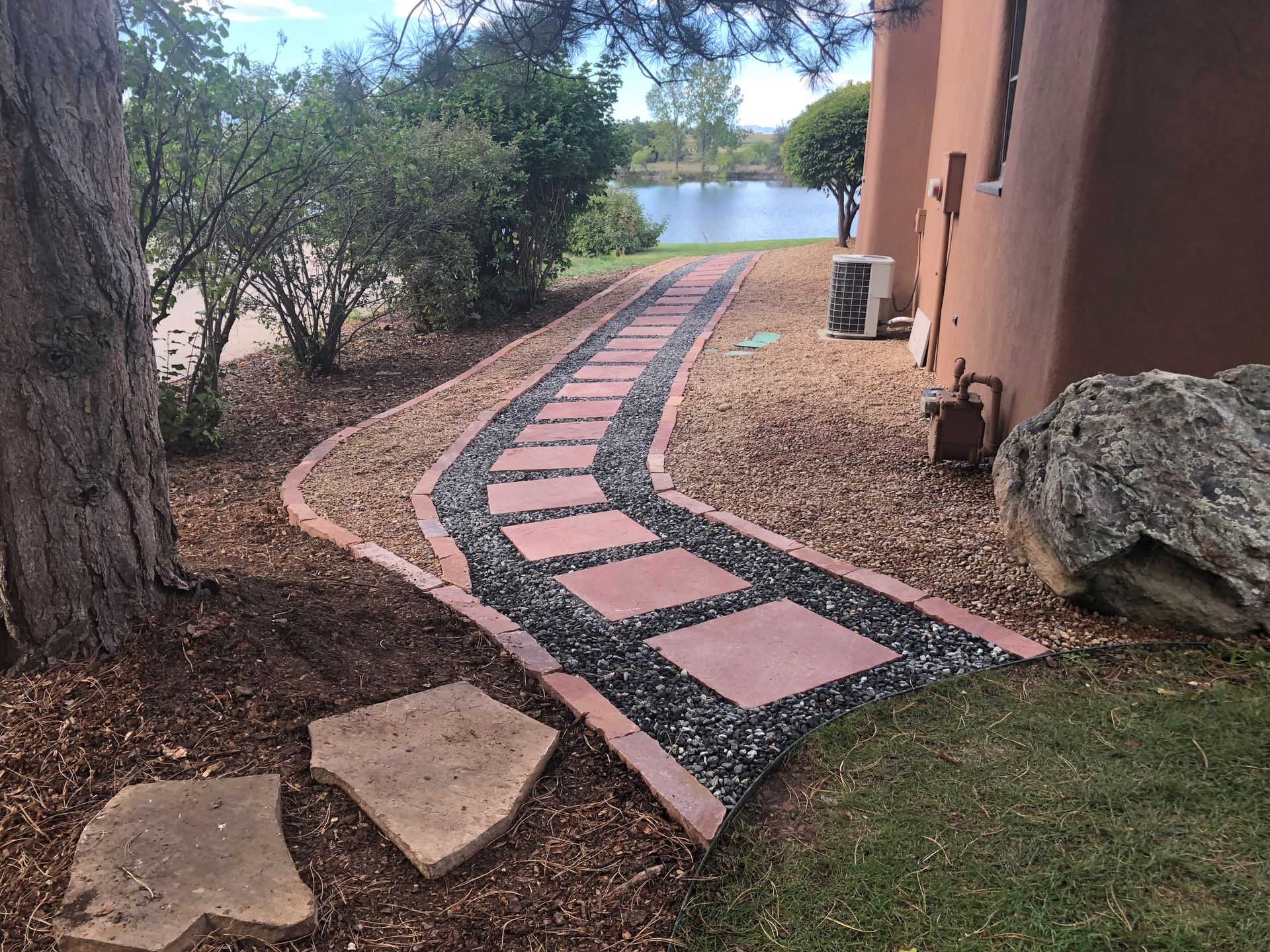 A stone pathway curves through a yard, next to a building and a body of water.