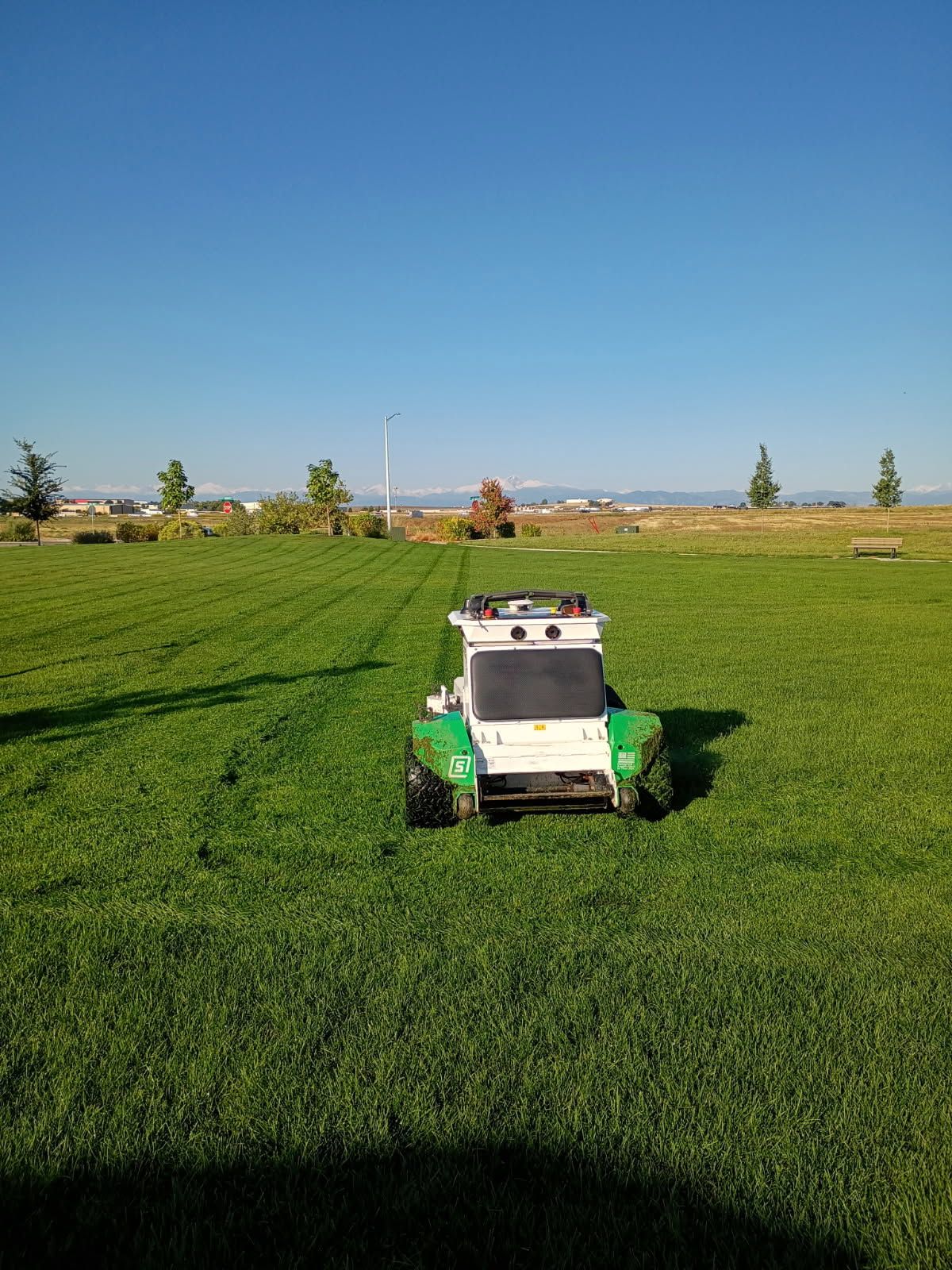 Robotic lawnmower, white and green, cutting grass in a sunny park.