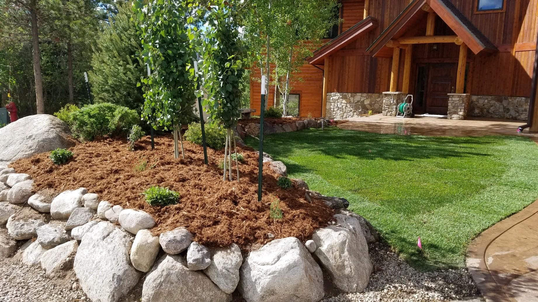 Landscaped yard with a stone retaining wall, mulch, trees, and a wooden house.
