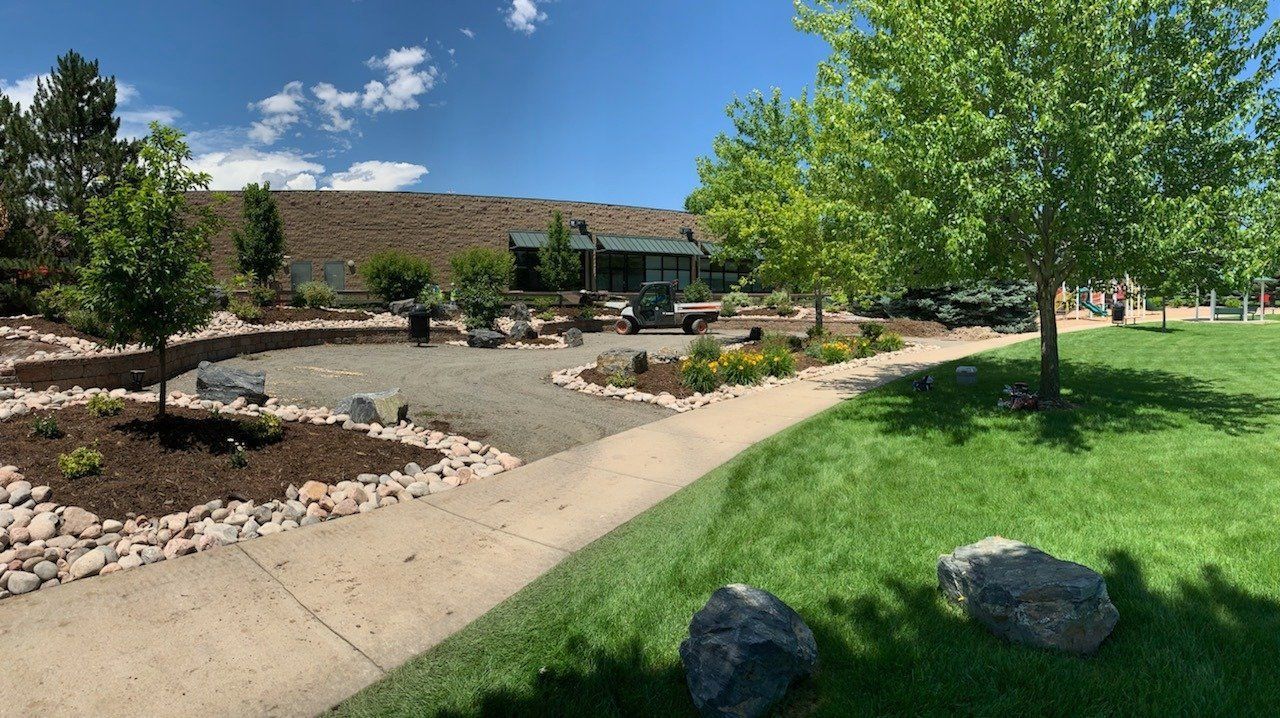 Park scene with a paved path, grass, trees, and a gravel seating area. A building is in the background, set against a hillside.