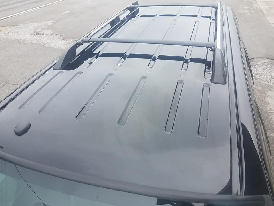 A high-angle view of a black car roof featuring a silver luggage rack against a paved outdoor background.