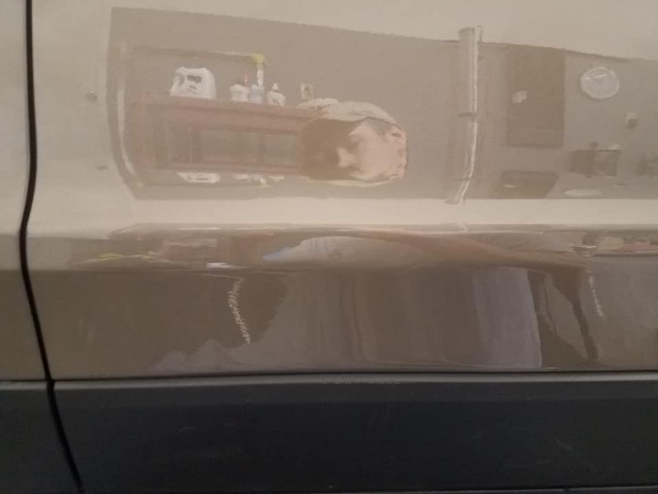 A reflection in a car door showing a person wearing a cap, with a garage shelf containing bottles visible above.
